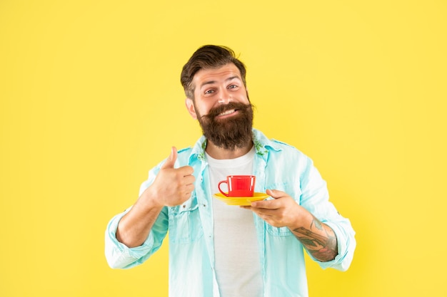 Chico barbudo feliz bebe café de la mañana muestra el pulgar hacia arriba en el café de fondo amarillo