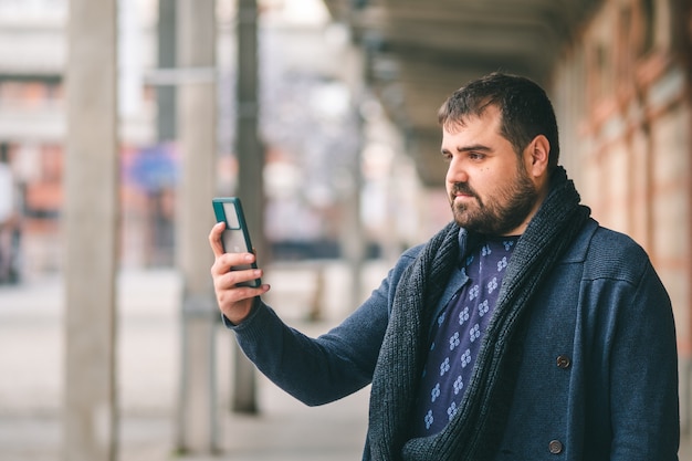 Chico barbudo en suéter azul de pie en la calle con su teléfono inteligente