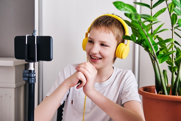 Chico blogger en auriculares amarillos hablando con seguidores Adolescente transmisión en vivo mirando a la pantalla del teléfono inteligente y gestos en casa