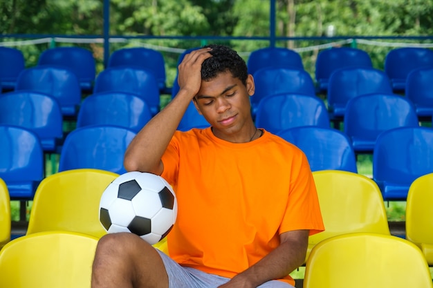 Chico brasileño con un balón de fútbol sentado en una tribuna de fútbol vacía