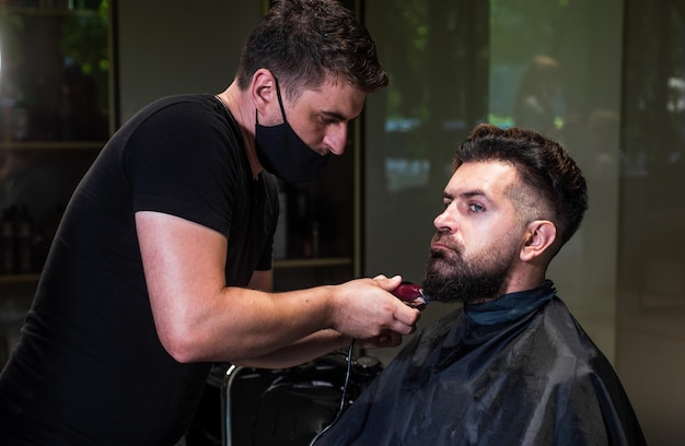 Chico brutal de aspecto mágico con maestro profesional de barbería uso de máquina de afeitar eléctrica hipster hombre barbero cuidado hombre barbudo en la silla de peluquería en el salón de belleza y peinado de moda y barba