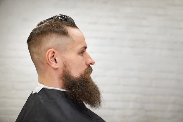 Chico brutal en la moderna peluquería. Peluquero hace peinado a un hombre con una larga barba. Maestro peluquero hace peinado con cortapelos