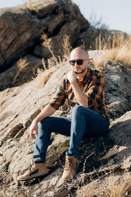 Chico calvo con barba en jeans camisa cálida y zapatos de trekking sobre rocas de granito