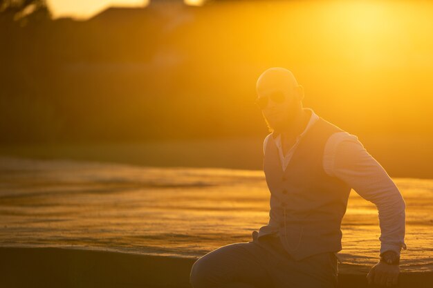 Foto chico calvo con una elegante barba y gafas de sol sobre un fondo borroso de la ciudad durante la espectacular puesta de sol. concepto de éxito y voluntad