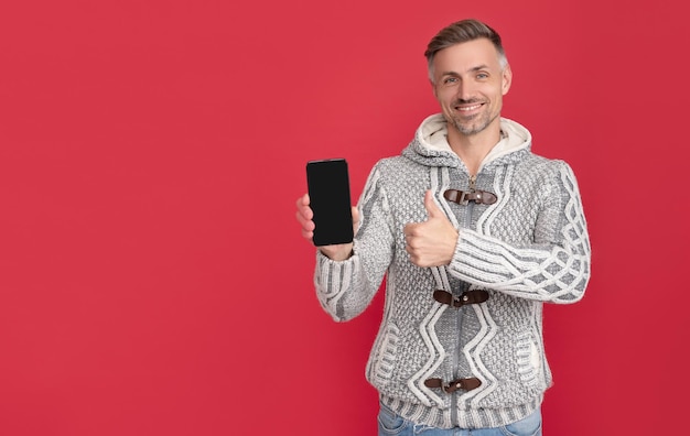 Chico canoso alegre en suéter que presenta un teléfono inteligente con espacio de copia en un anuncio de fondo rojo