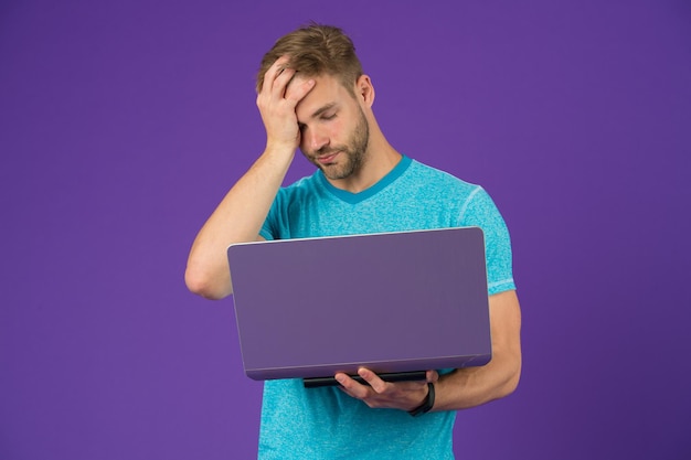Chico cansado con computadora sobre fondo violeta. El hombre con camiseta azul trabaja en una laptop con fondo morado. Navegación por Internet y mundo virtual. Blog o blogs y weblog. La vida moderna en la red social.