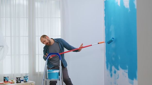 Chico cantando durante la reparación del hogar con un cepillo de rodillo bañado en pintura azul. Hipster renovando su casa. Redecoración de apartamentos y construcción de viviendas mientras se renueva y mejora. Reparación y decoración