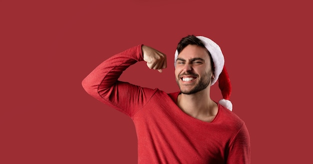 Chico caucásico guapo joven en suéter rojo y sombreros de Santa se encuentra sobre fondo rojo en el estudio y muestra el poder de los bíceps. Cerrar retrato chico europeo con humor navideño. Banner de vacaciones. Año nuevo.