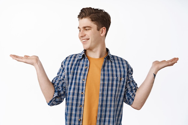 Chico caucásico joven que muestra las manos abiertas, sosteniendo el artículo y mirando la palma con un lugar vacío para sus productos, sonriendo complacido, comprando, de pie sobre blanco