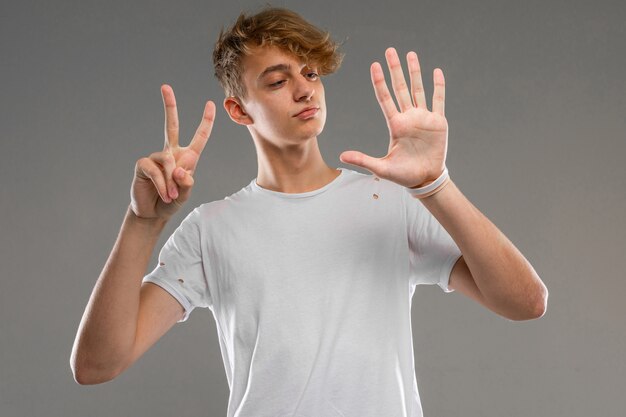 Chico caucásico sonriente con una camiseta blanca muestra siete dedos en una pared gris