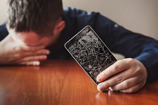 Chico con cerdas, está mostrando su mano en un teléfono inteligente. Pantalla rota del teléfono sin marco moderno. chico llorando con los ojos cerrados y las manos en la cara.