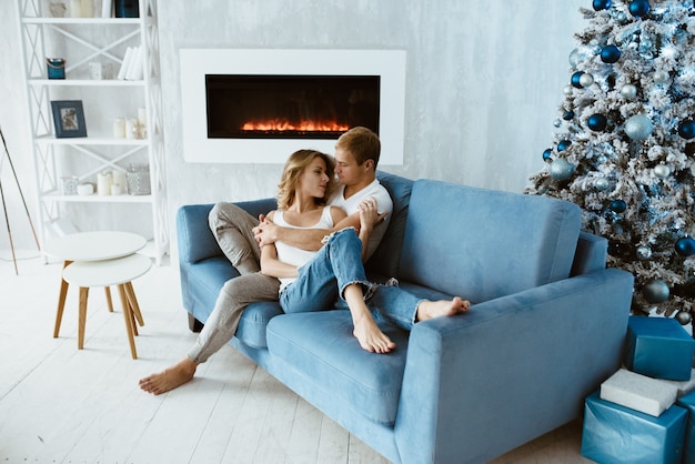 El chico y la chica se abrazan y besan en el sofá azul. Árbol de Navidad decorado con juguetes. Chimenea electronica.