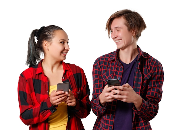 Un chico y una chica con camisas a cuadros están uno al lado del otro y se miran con una sonrisa, sosteniendo sus teléfonos inteligentes en sus manos aisladas sobre fondo blanco