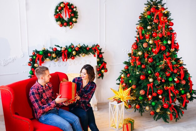 El chico y la chica están sentados en el sofá de la habitación con chimenea y árbol de Navidad. el chico le da un regalo a la chica