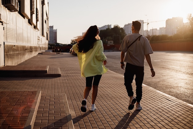 Chico y chica con estilo joven caminan juntos en la plaza para aparcar junto al edificio en la puesta de sol.