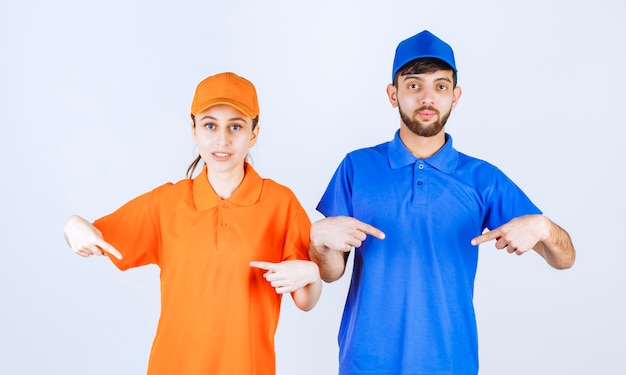 Foto chico y chica mensajero con uniformes azules y amarillos apuntando a sí mismos y sintiéndose emocionados.