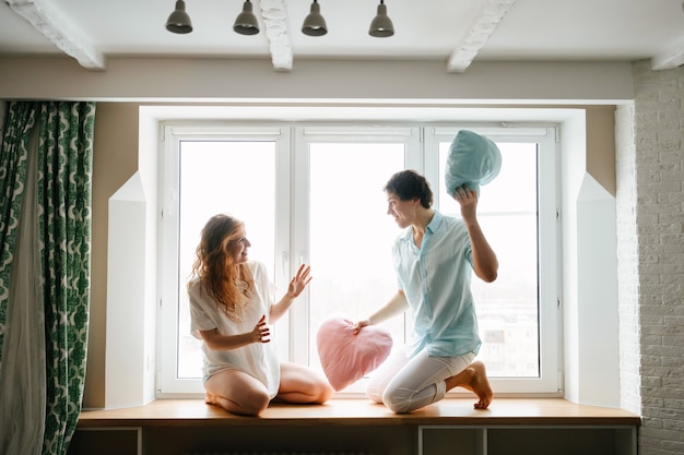 Chico y chica de pareja juegan con almohadas junto a la ventana. Ropa blanca y azul. Día de San Valentín.