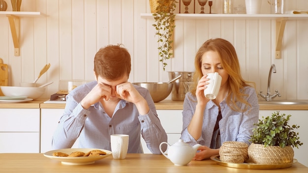 Chico y chica rubia pareja triste desayunar bebiendo té con galletas en silencio y el hombre bosteza en la mesa en la cocina, luz del sol