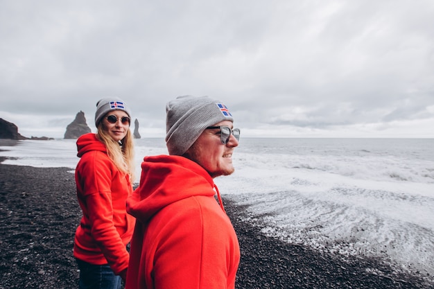 Un chico y una chica con suéteres rojos y sombreros grises se abrazan en una playa negra, una feliz pareja sonriente elegante caminando y besándose en Islandia