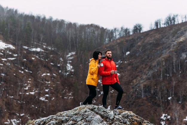 Chico y chica viajan en las montañas. El hombre y la mujer beben té caliente en las montañas. Una pareja de enamorados disfruta del té caliente. Escalando la montaña.