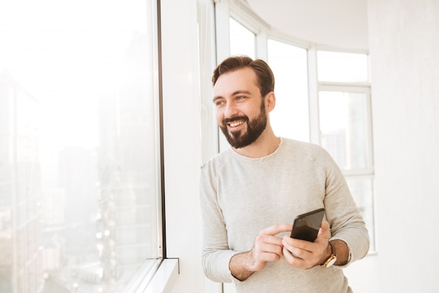 Chico comunicativo con camisa blanca, mirando por una ventana grande, mientras chateas o escribes mensajes de texto en el teléfono móvil