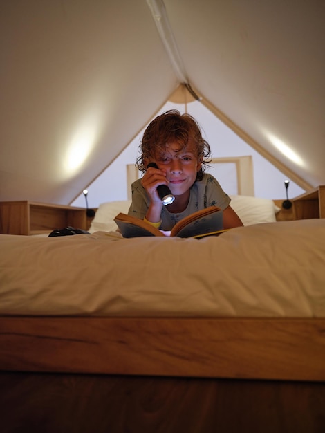 Chico contento brillando con una linterna en el libro y mirando a la cámara mientras está acostado en una cómoda cama blanca en una mansarda en casa