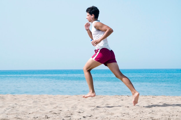 Chico corriendo con auriculares y una botella de agua en la playa. Joven corriendo en el lado del mar.