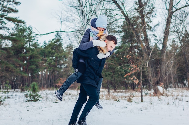 Chico dando a su novia a cuestas en el bosque de invierno. Joven pareja amorosa divirtiéndose al aire libre