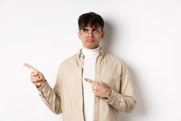 Chico decepcionado con gafas mirando y apuntando a la izquierda, enfurruñado disgustado, quejándose de una mala oferta, de pie molesto en la pared blanca.