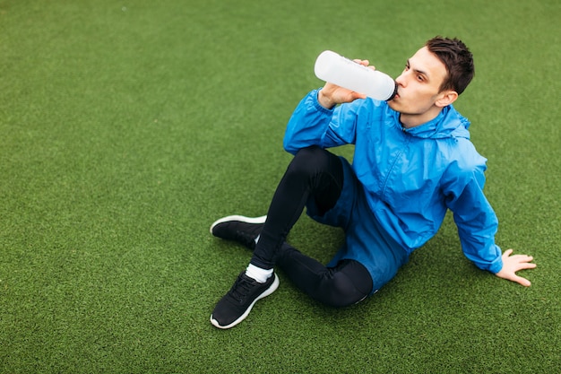 Chico después del ejercicio, beber agua en el campo de fútbol. Retrato de hombre hermoso en ropa deportiva.