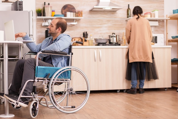 Chico con discapacidad para caminar en silla de ruedas usando laptop en cocina y esposa está cocinando comida. Hombre discapacitado paralítico discapacitado con discapacidad para caminar que se integra después de un accidente.