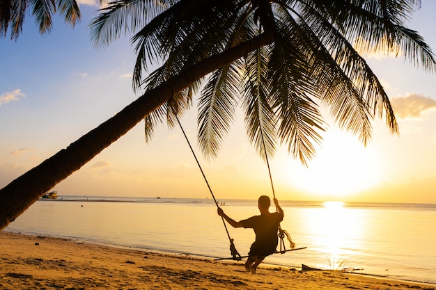 El chico disfruta del atardecer montando en un columpio en la playa tropical. Siluetas de un chico en un columpio colgado de una palmera, viendo la puesta de sol en el agua.