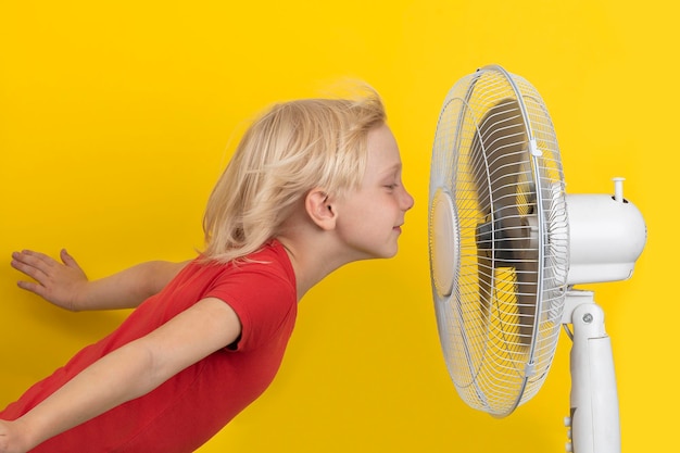 Chico disfrutando del aire fresco. Niño y ventilador sobre fondo amarillo. Regulación de la temperatura ambiente.