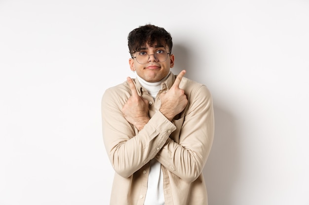 Chico divertido confundido con gafas apuntando con el dedo a un lado, mostrando dos variantes y aspecto indeciso, no puedo elegir, de pie sobre fondo blanco.