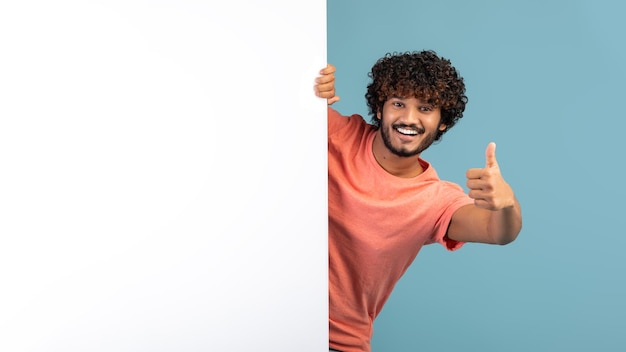 Foto chico eatsern positivo de pie junto a un tablero de publicidad blanco gesticulando