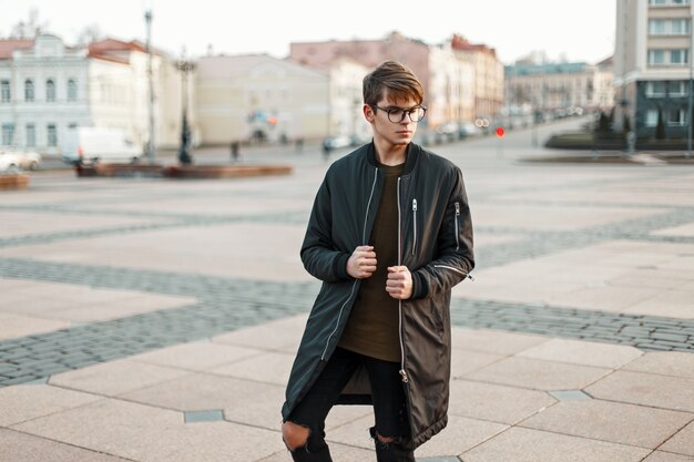 Chico elegante con gafas en chaqueta de moda y jeans rotos con zapatillas negras en la ciudad