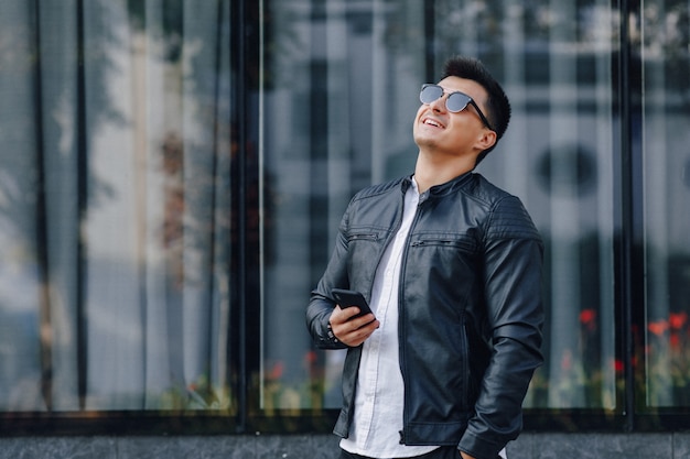 Chico elegante joven con gafas en chaqueta de cuero negro con teléfono sobre fondo de cristal