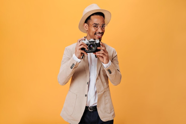 Chico elegante con sombrero sostiene una cámara retro sobre fondo naranja Hombre fresco con chaqueta beige tomando fotos y sonriendo en aislado