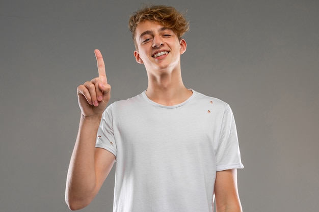 Chico emocional guapo posando en estudio contra gris, chico con una camiseta gris muestra el pulgar hacia arriba