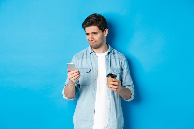 Chico escéptico y decepcionado leyendo un mensaje molesto en el teléfono, sosteniendo una taza de café, de pie sobre una pared azul