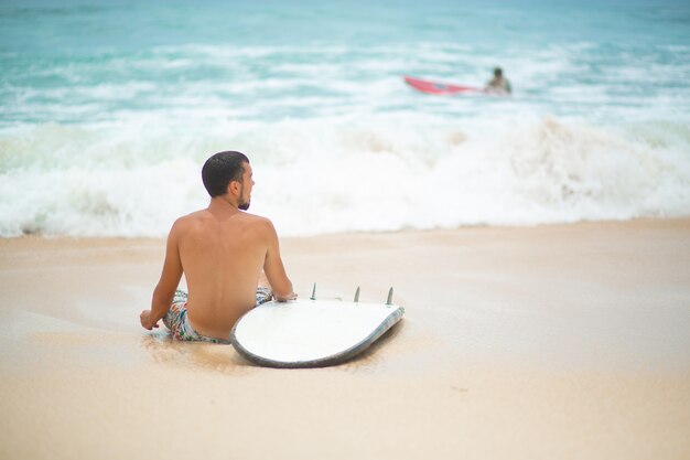 El chico está descansando en una playa tropical de arena, después de surfear.