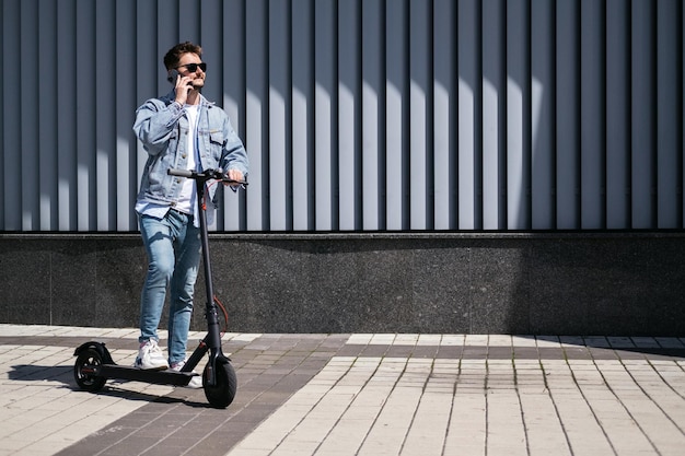 El chico está montando un scooter eléctrico contra el fondo de la ciudad. U cantar teléfono.