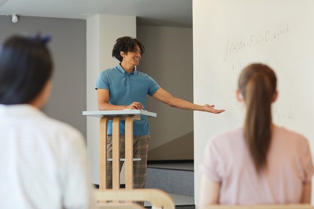 Foto chico estudiante alegre en traje casual gesticulando con la mano mientras presenta su investigación en la clase universitaria