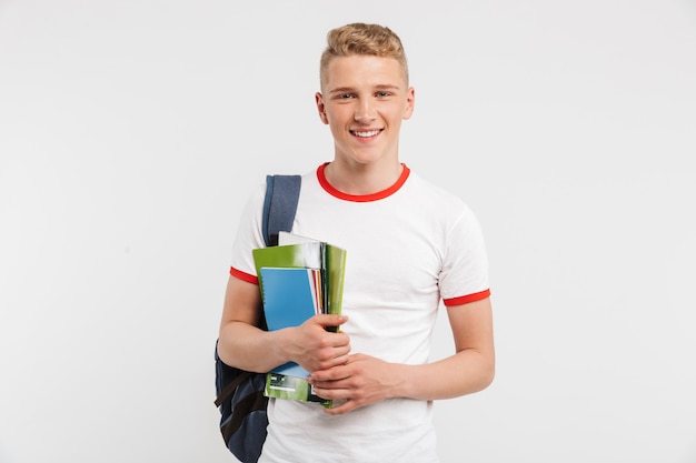 Chico estudiante europeo con mochila sonriendo y posando con coloridos libros de ejercicios en manos aisladas en blanco