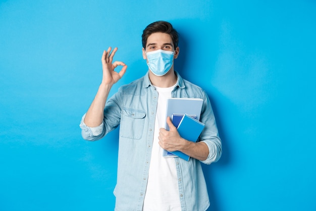 Chico estudiante en máscara médica mirando feliz, sosteniendo cuadernos, mostrando el signo de ok, de pie sobre la pared azul