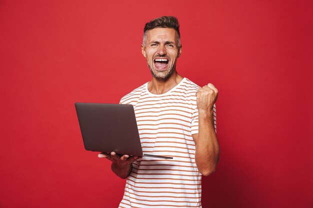 Chico exitoso en camiseta a rayas sonriendo y sosteniendo portátil gris aislado en rojo