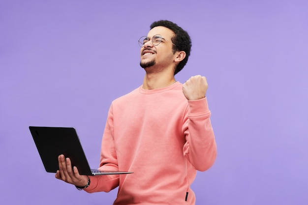 Chico extasiado en ropa casual sosteniendo una computadora portátil y expresando entusiasmo