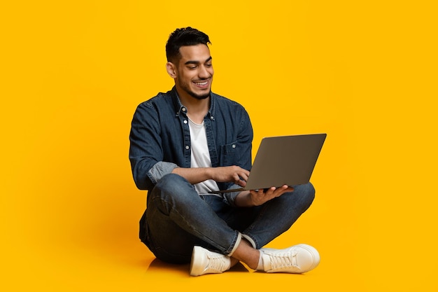 Chico feliz del Medio Oriente usando un cuaderno moderno aislado en amarillo