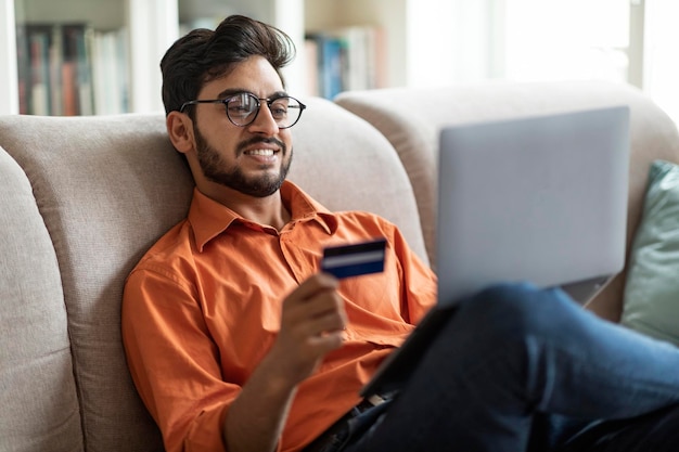 Chico feliz del medio oriente usando tarjeta de crédito y computadora portátil