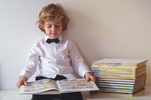 Chico feliz en ropa formal con corbata de papagaio leyendo un libro interesante mientras está sentado en la mesa cerca del fondo blanco en la habitación